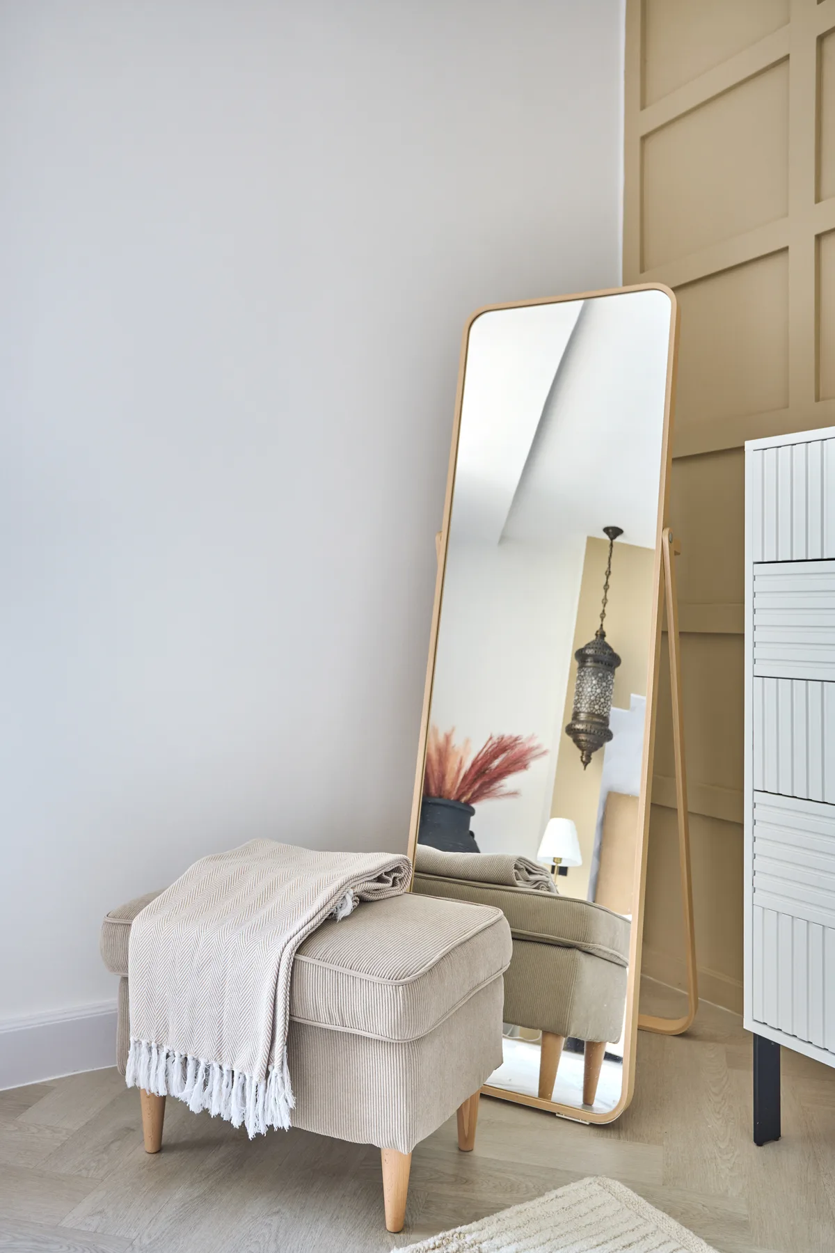 Un otomán tapizado de color claro está junto a un espejo alto apoyado en una habitación luminosa.