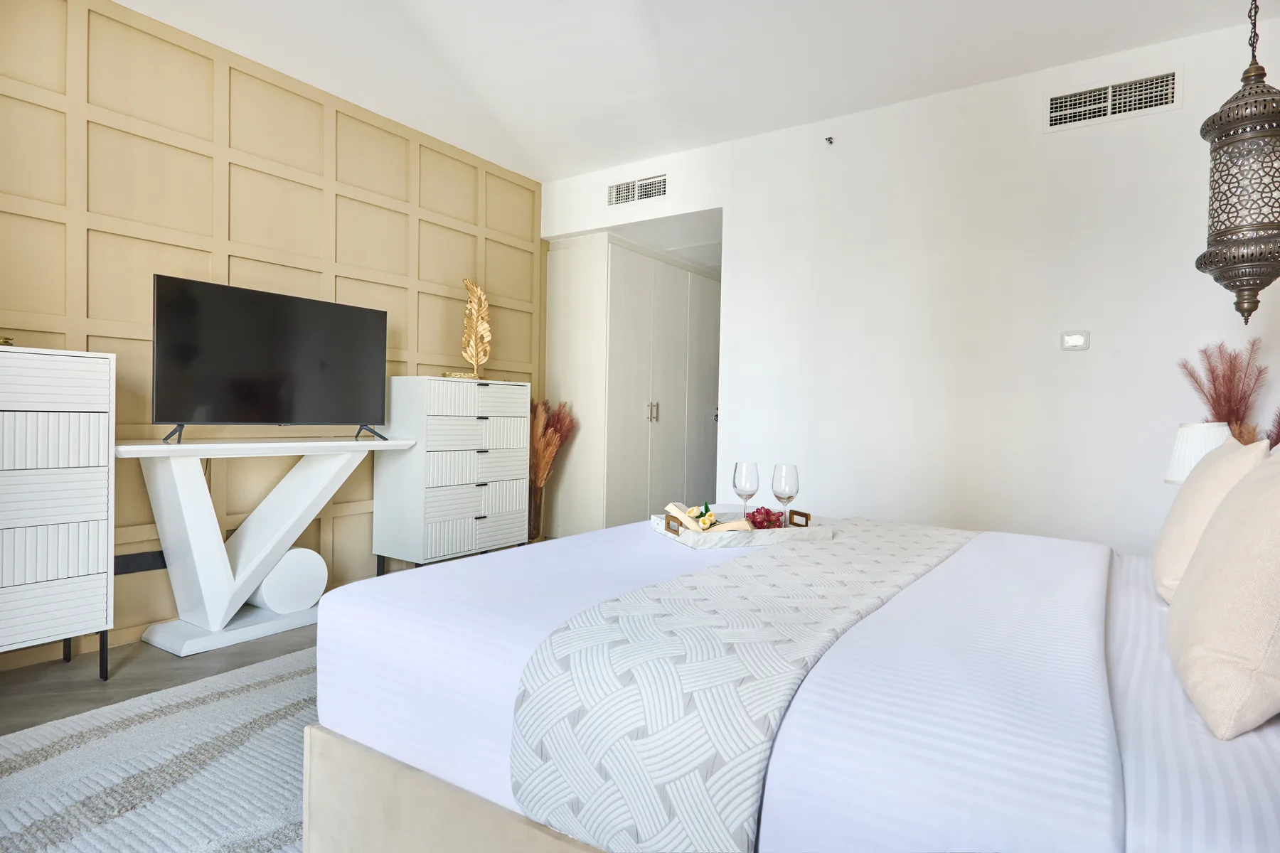 Un dormitorio luminoso con una cama tendida y un televisor frente a una pared panelada.