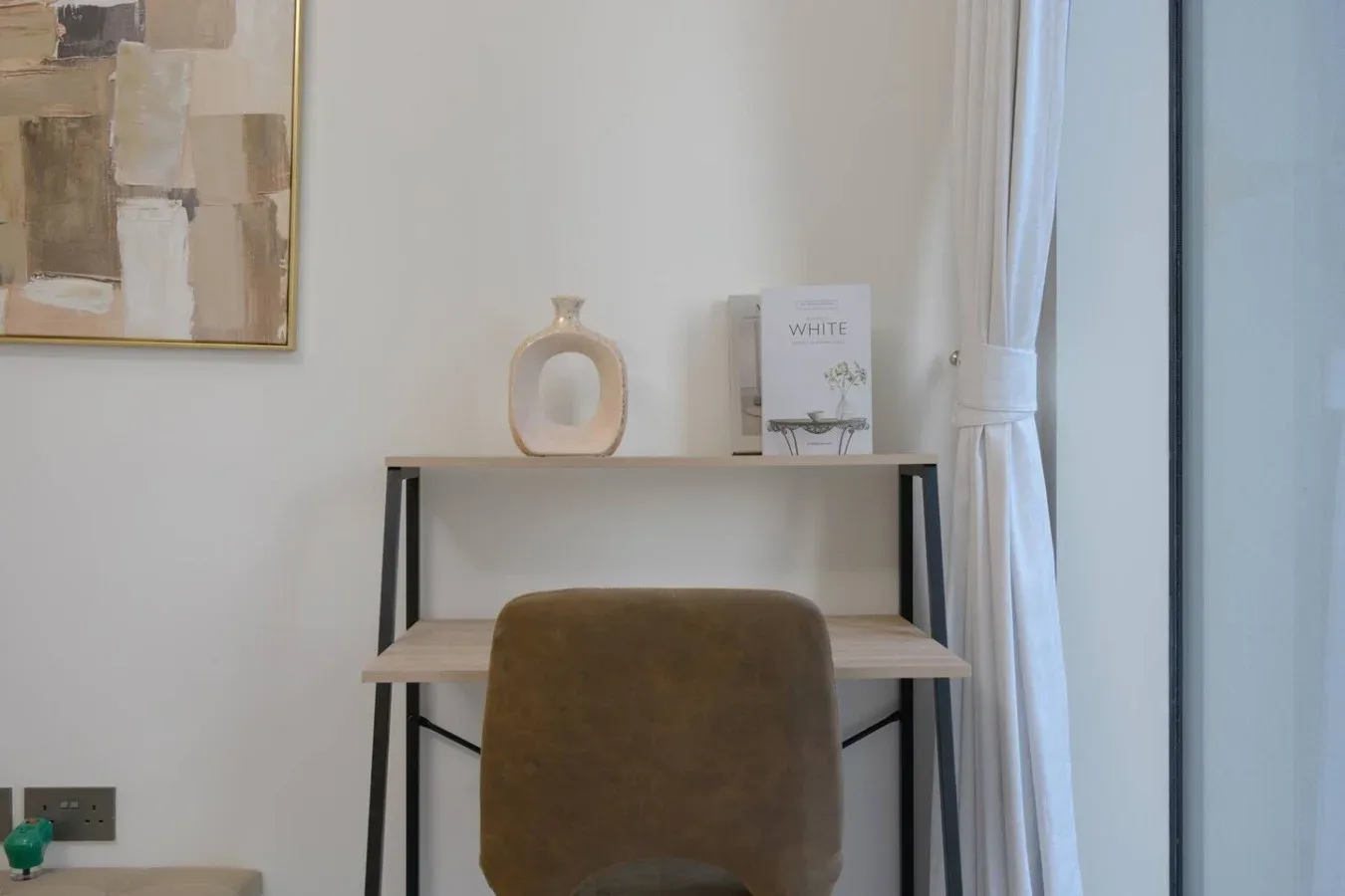 Un escritorio pequeño con una silla está bajo una obra enmarcada junto a una ventana con cortina.