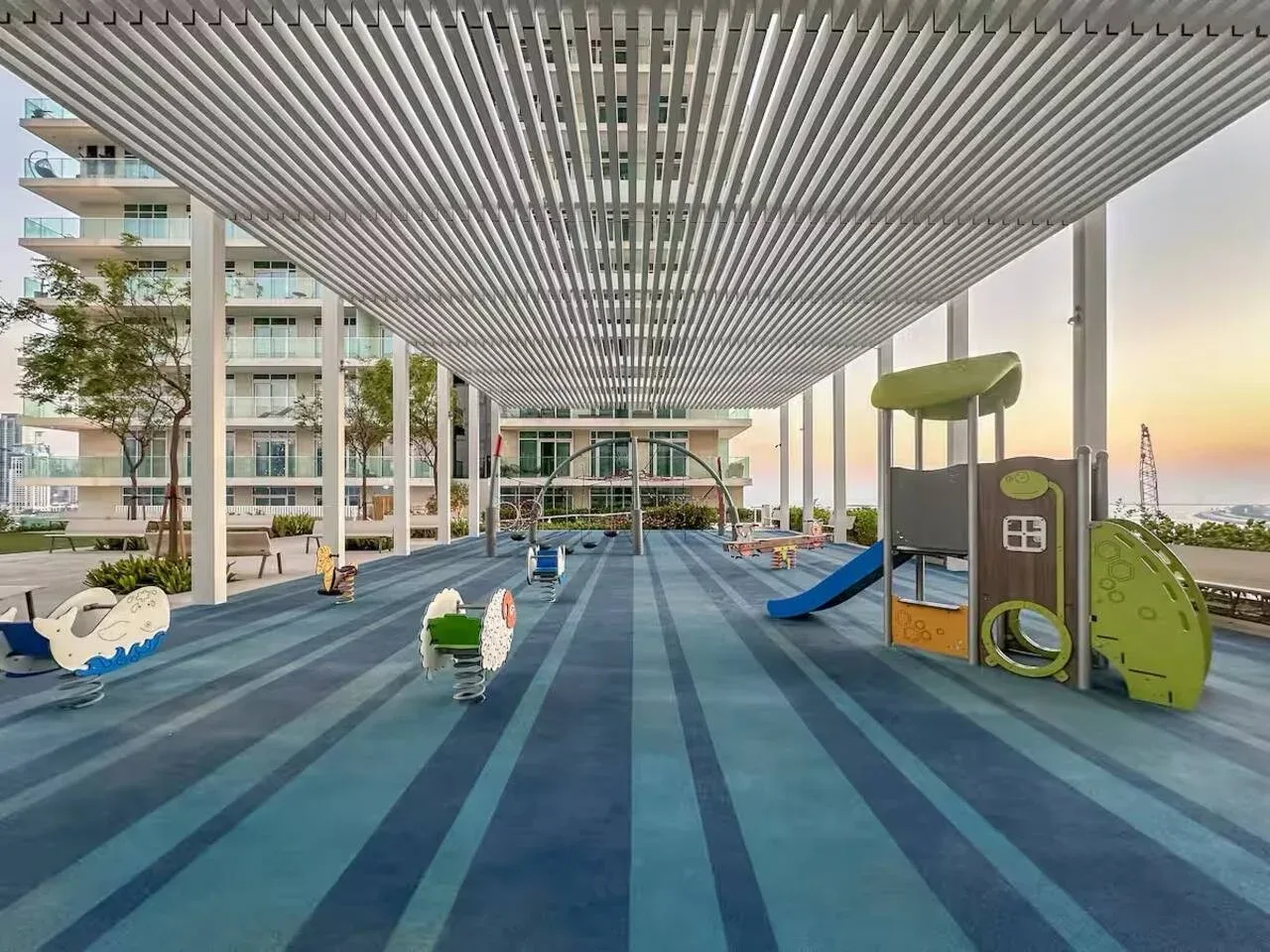 Un parque infantil exterior cubierto, con juegos coloridos y edificios de apartamentos al fondo.