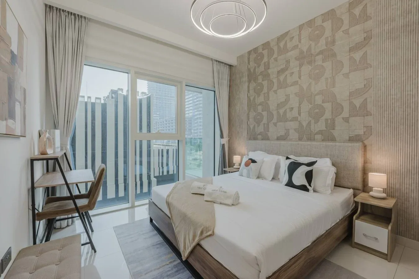 Un dormitorio con una cama, mesitas de noche y grandes ventanales con edificios a la vista.