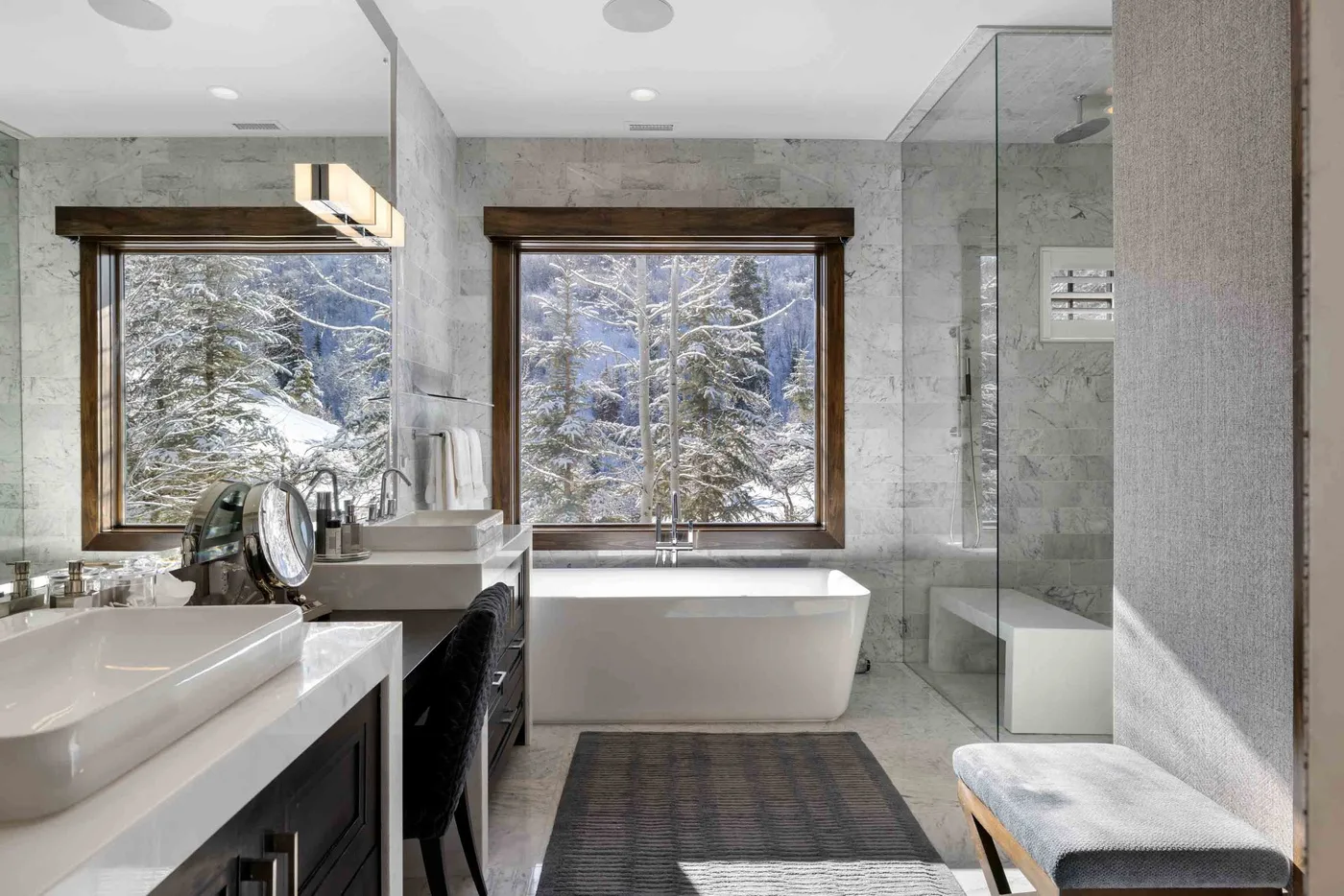 A modern bathroom has a bathtub, glass shower, and large windows overlooking snowy trees.