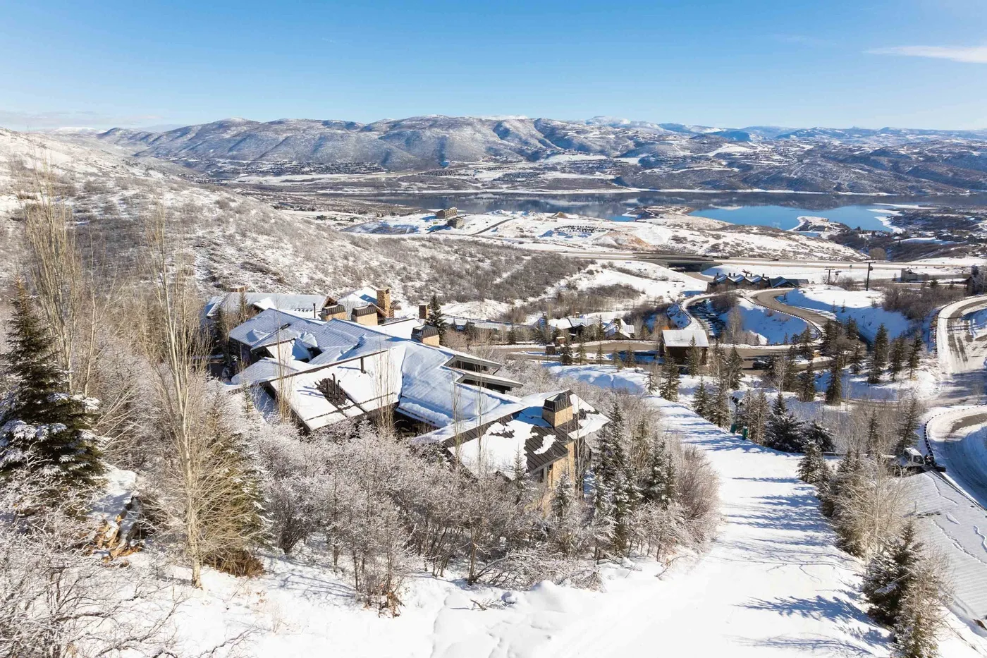 Snow-covered buildings sit among trees on a hillside overlooking a lake and mountain landscape.