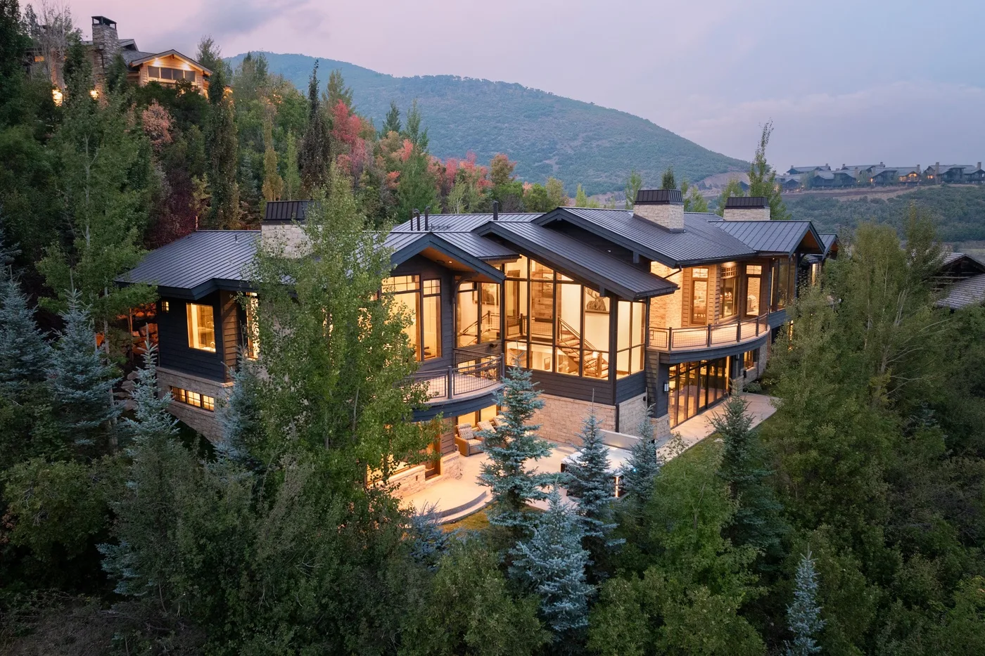 A large illuminated house sits among trees, with a mountain ridge in the background.