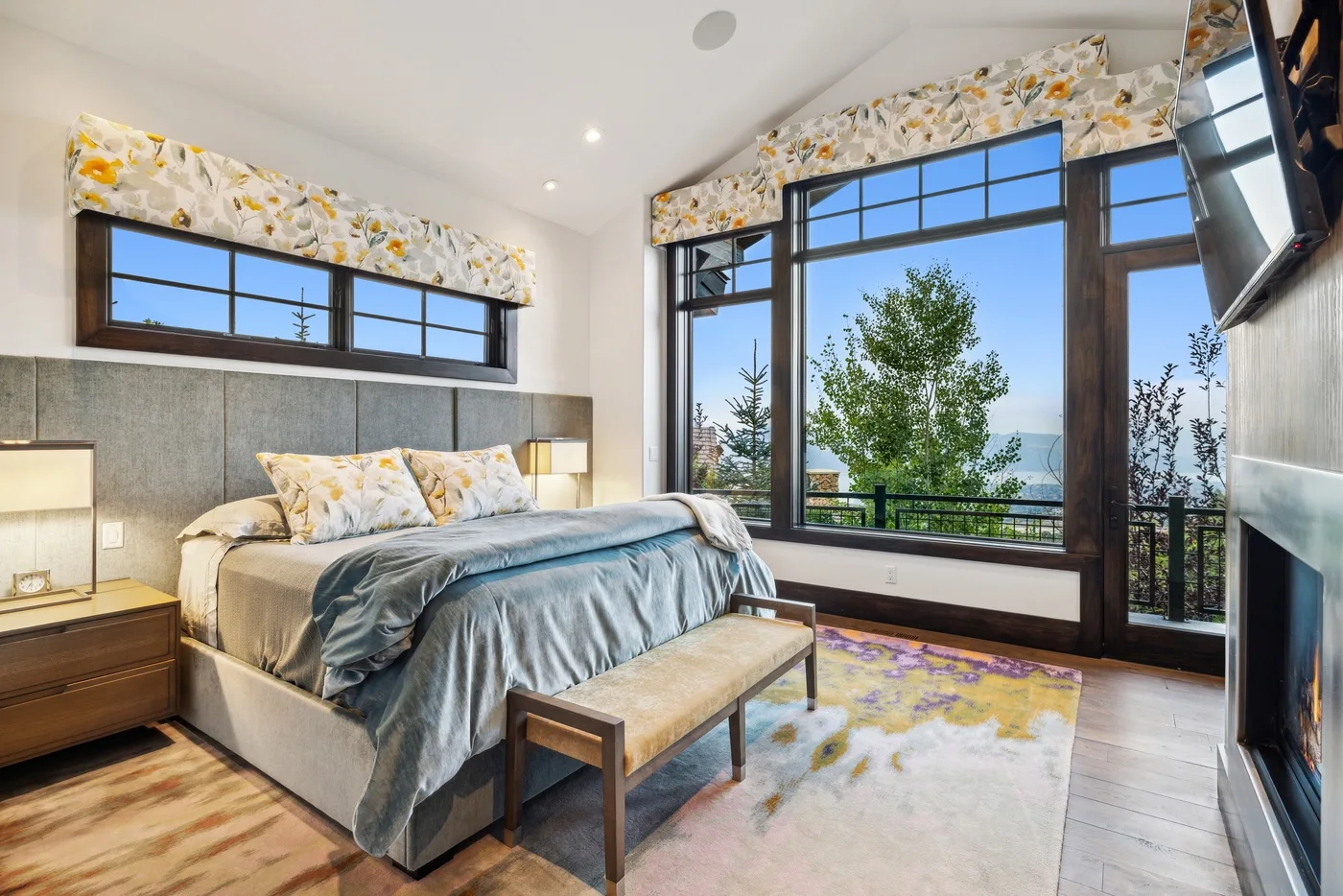 A bright bedroom with a bed beside large windows overlooking trees.