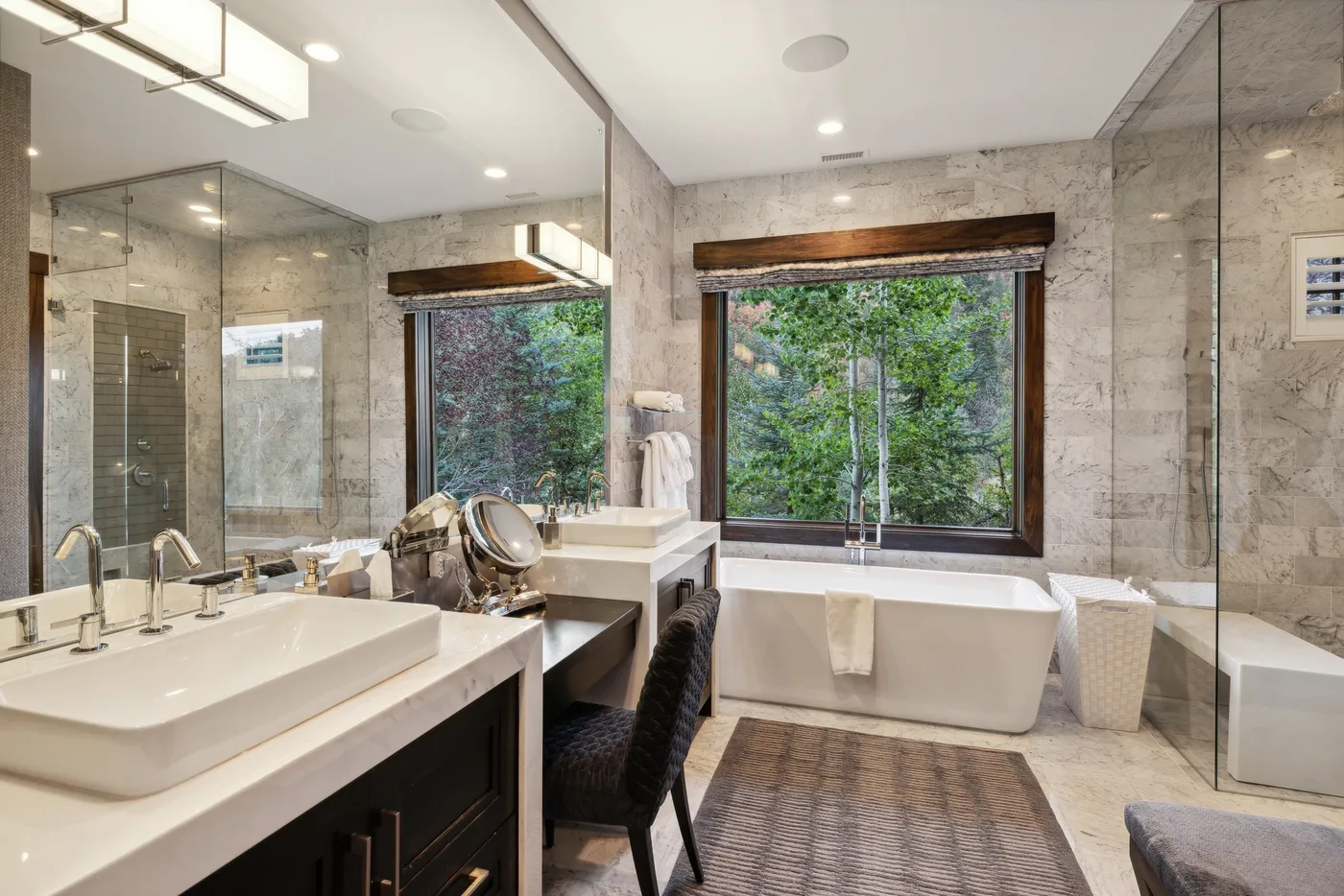 A marble bathroom with a bathtub, double sinks, and large windows overlooking greenery.