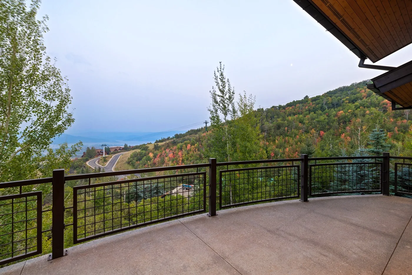 A terrace with a dark railing overlooks a winding road and wooded hills.