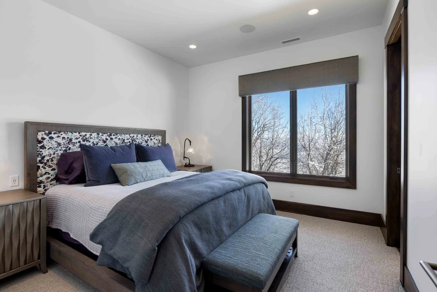 A bedroom with a bed, bench, and large window overlooking trees.