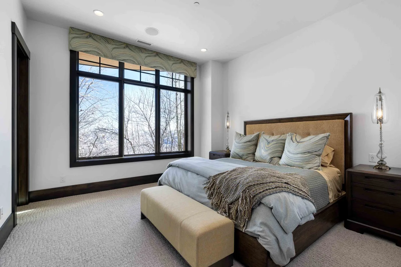 A bedroom with a large bed, upholstered bench, and wide window.