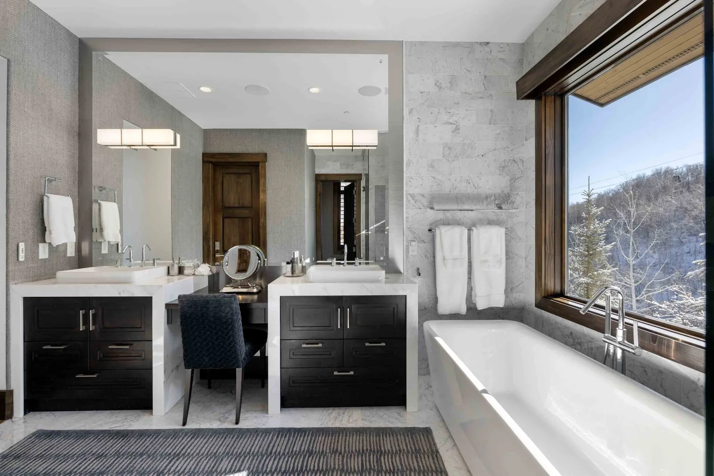 A modern bathroom with twin vanities and a bathtub beside a large window.