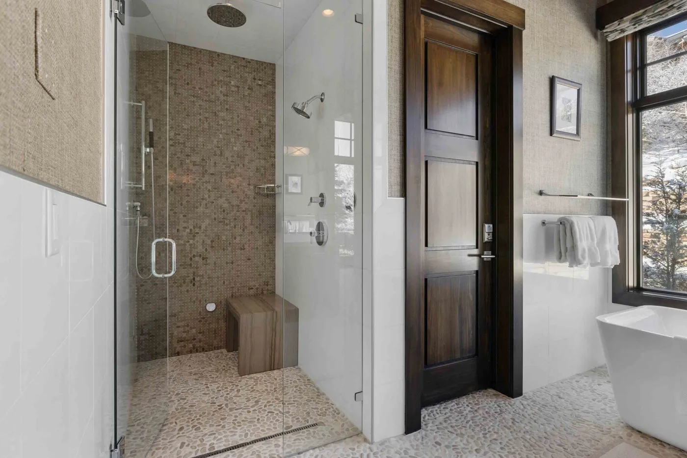 A bright bathroom with a glass shower, dark wood door, and freestanding bathtub.