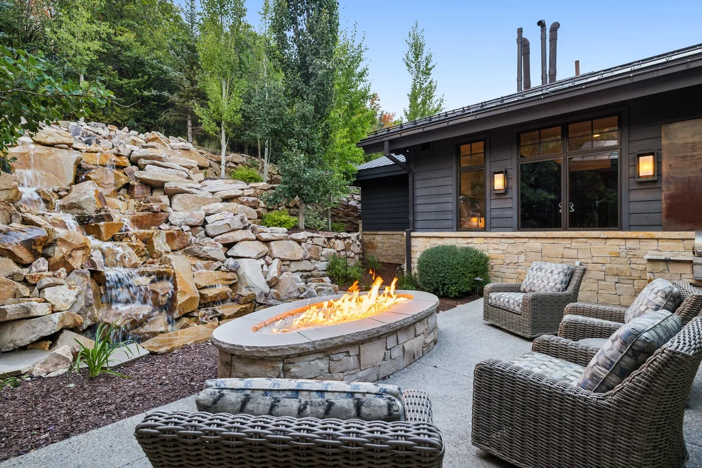 An outdoor patio features a stone fire pit, woven seating, and a rocky garden slope.