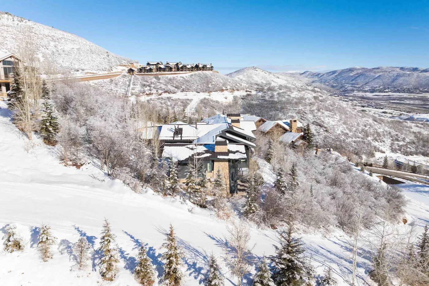 Snow-covered hillside homes overlook a broad mountain valley under a clear blue sky.