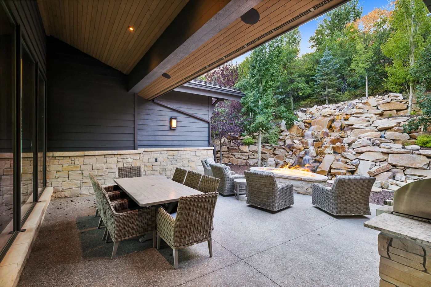 A covered outdoor patio with seating and a stone fire feature beside a wooded hillside.