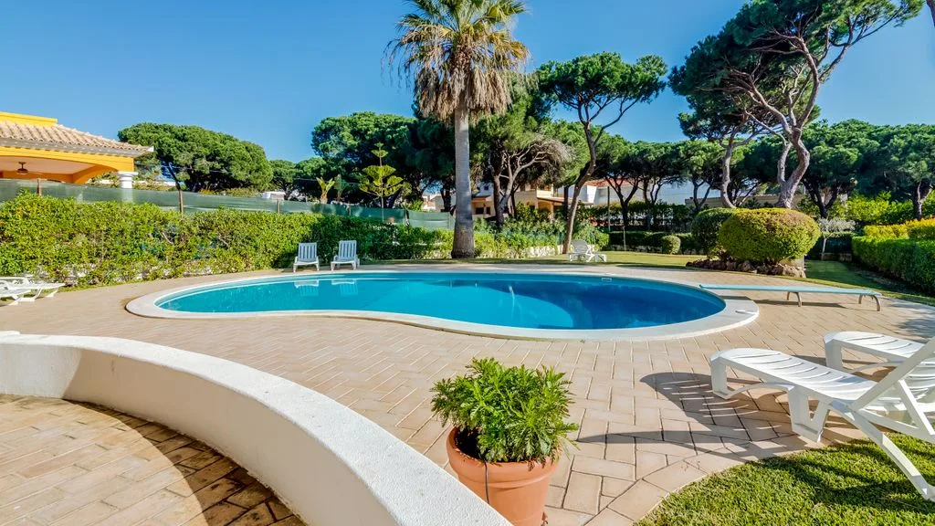 An outdoor swimming pool surrounded by tiled paving, lounge chairs, and trees.