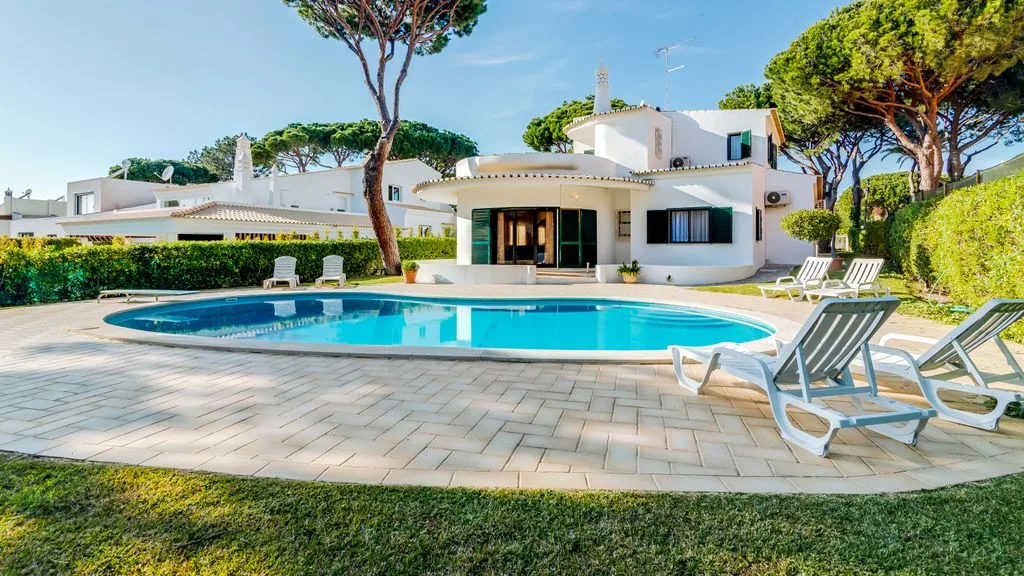 A modern white villa stands behind a swimming pool and paved patio.