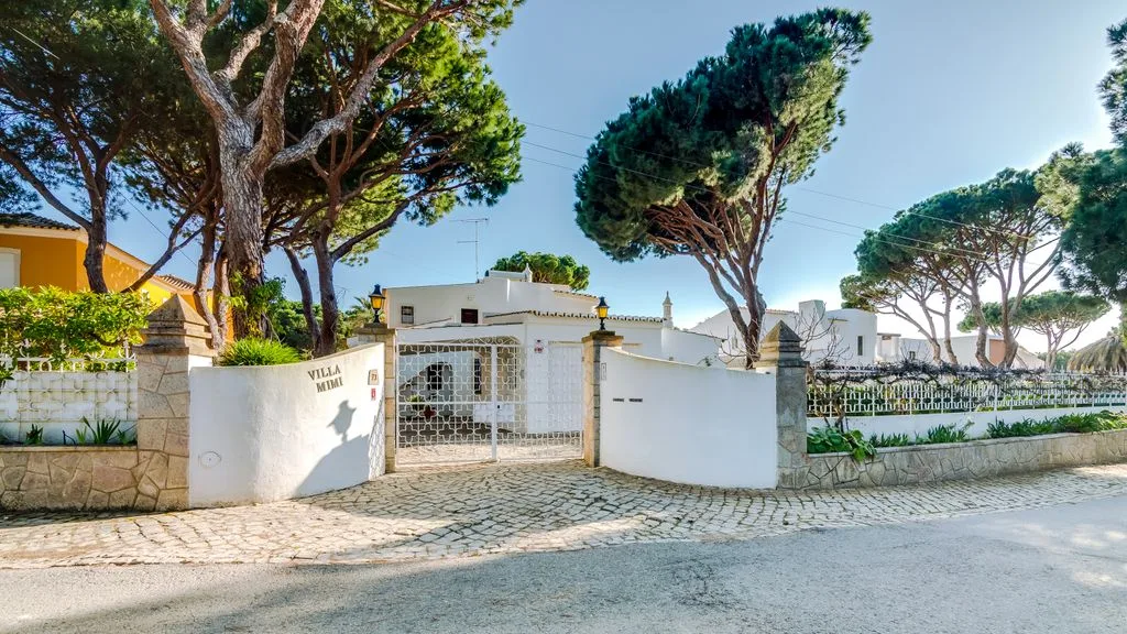 A white villa entrance with a gated driveway and tall trees.