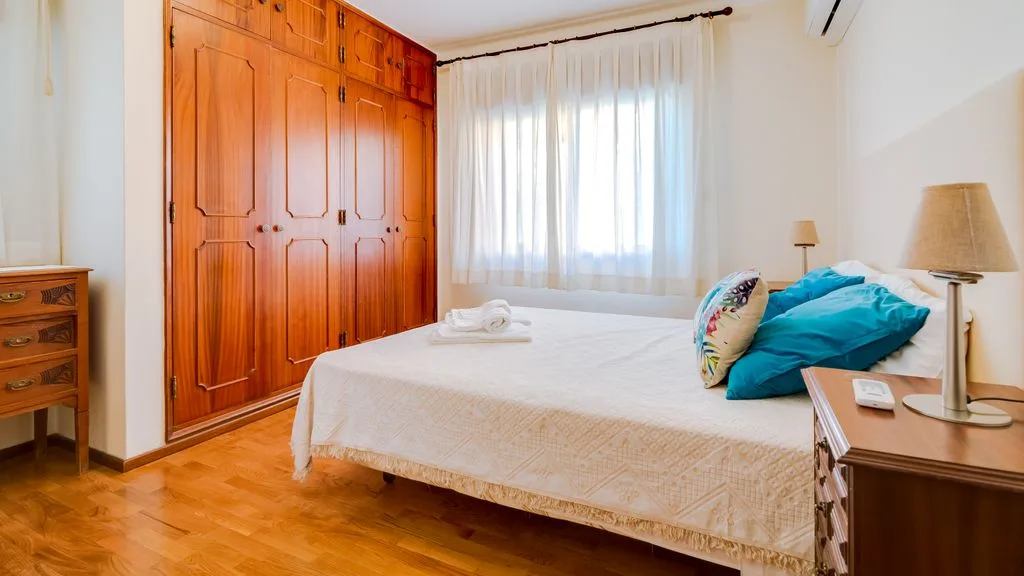 A bedroom with a large wooden wardrobe and a bed beside a window.