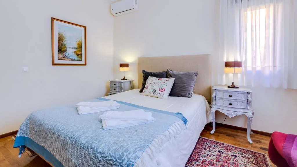A bedroom with a neatly made bed, bedside tables, and a window with sheer curtains.