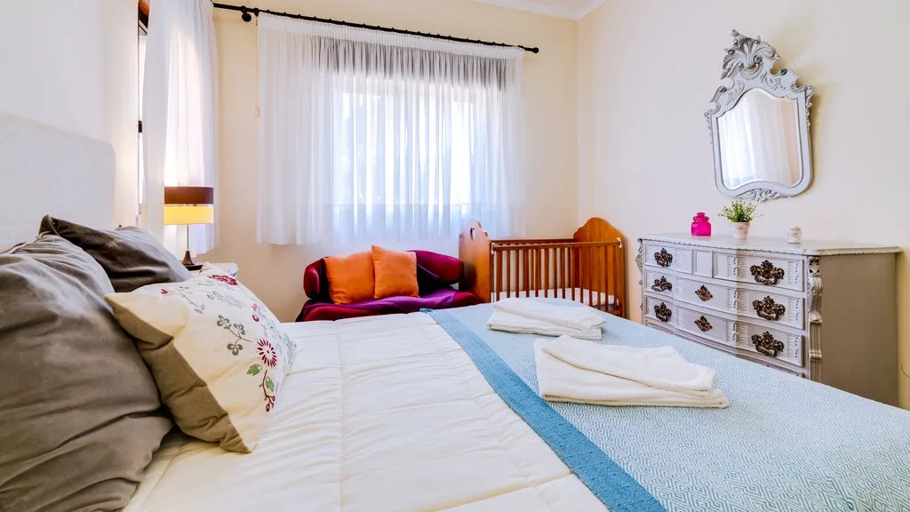A bright bedroom with a bed, a crib, and a dresser beneath a window.