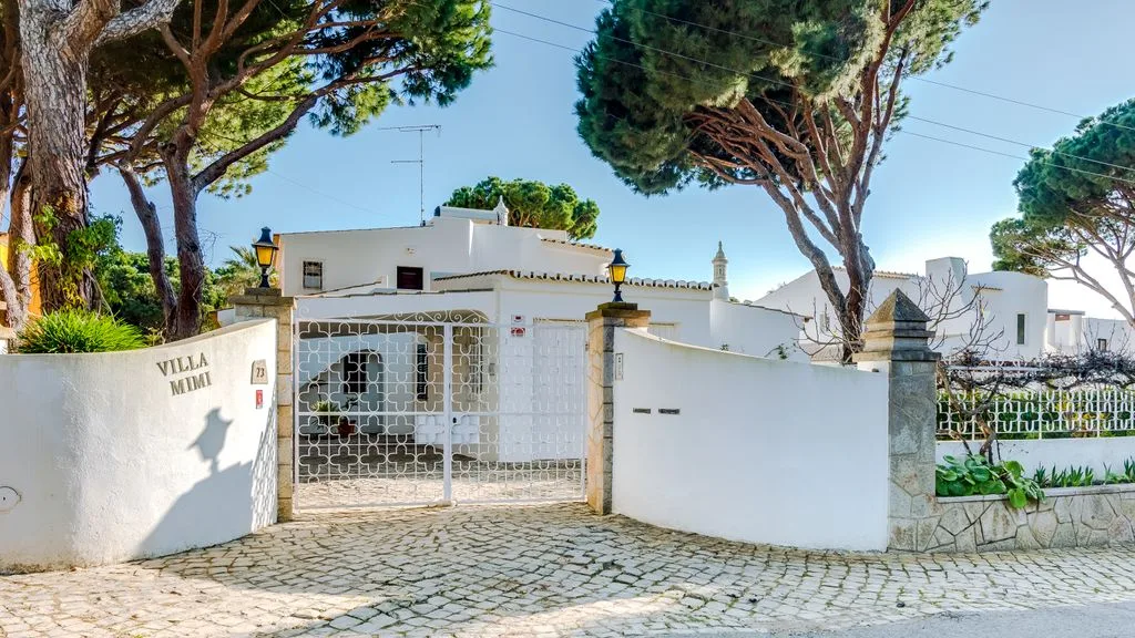 A white villa entrance with a metal gate beneath tall trees.