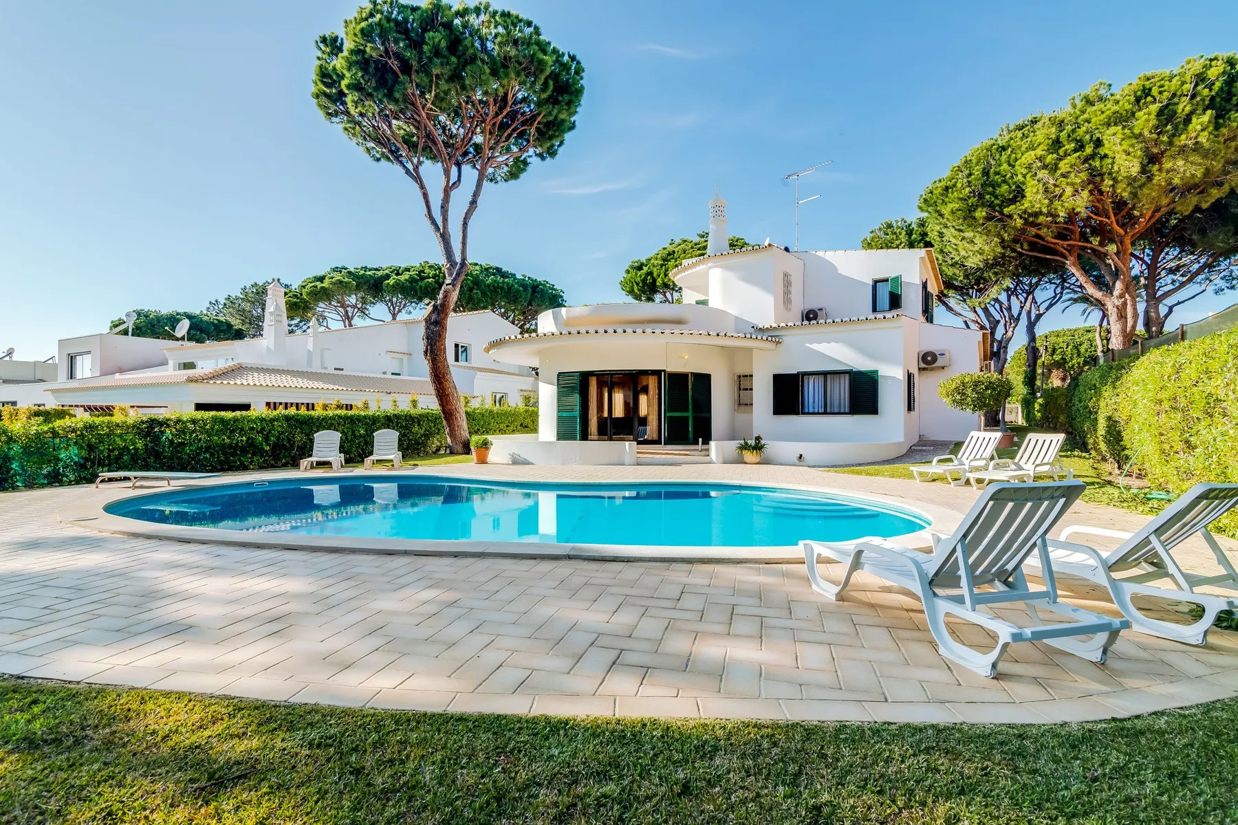 A white house overlooks a swimming pool, with lounge chairs on the paved patio.