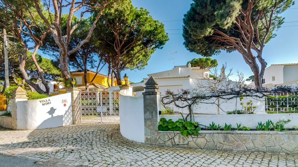 A gated property entrance with white walls, trees, and a cobblestone driveway.