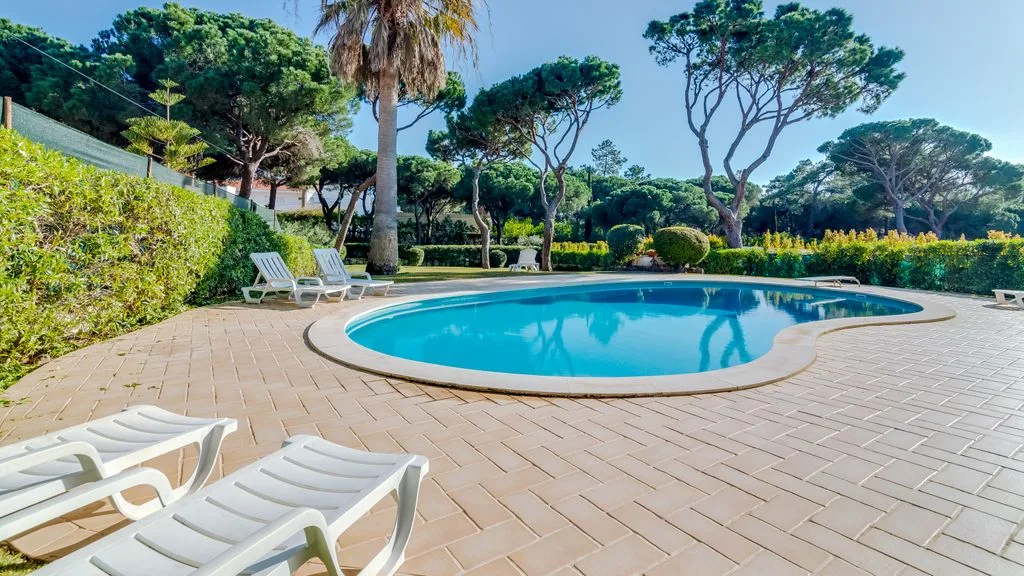 An outdoor swimming pool sits beside a paved deck and leafy trees.