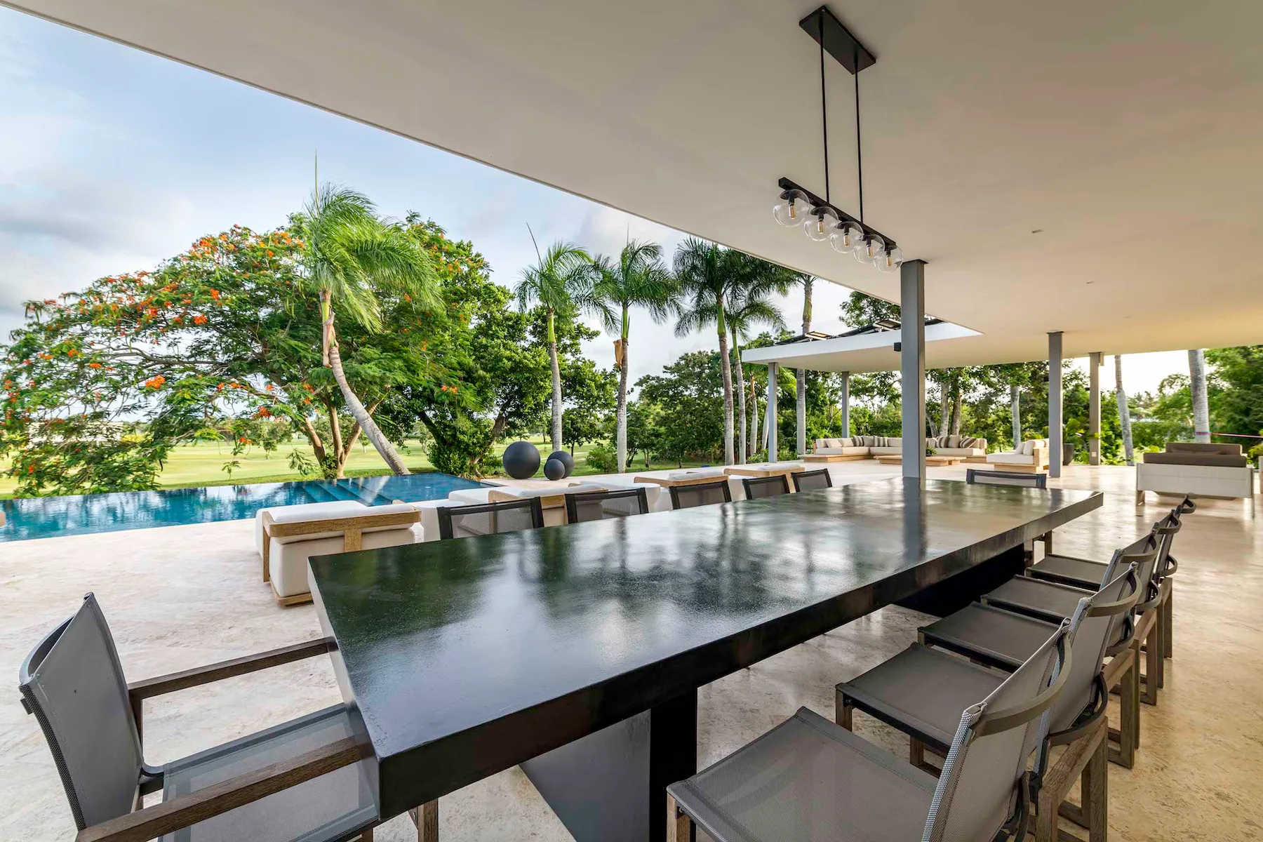 Comedor exterior cubierto con una mesa larga y vistas a una piscina y palmeras.