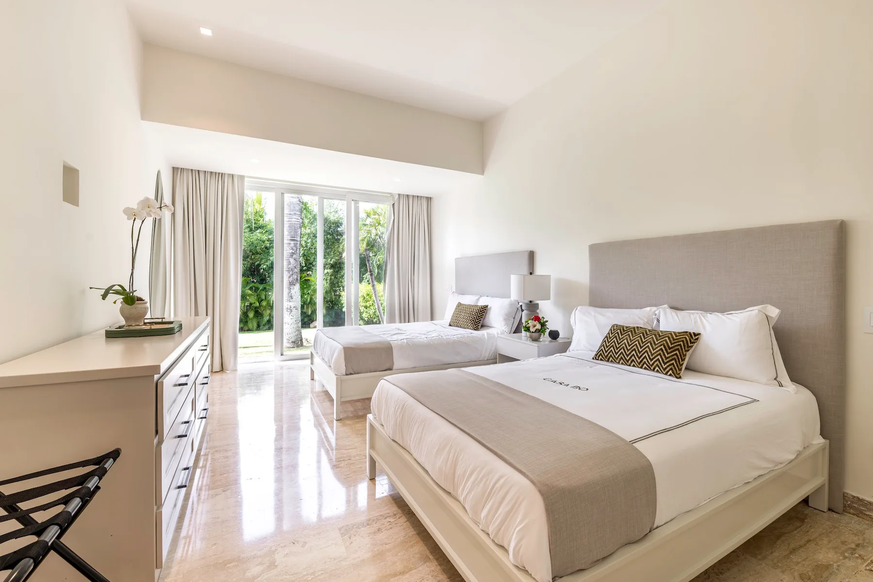 Un dormitorio luminoso con dos camas, lámparas de noche y puertas de cristal junto a la vegetación.