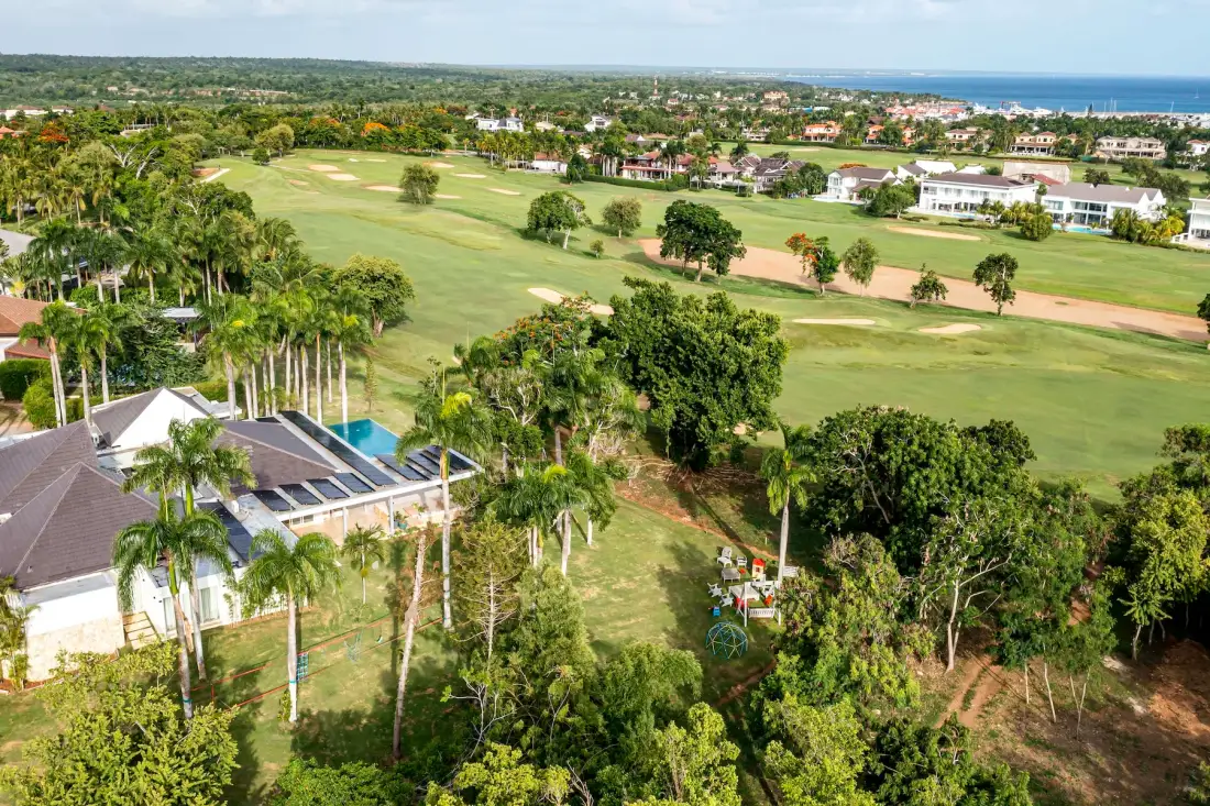 Vista aérea de un campo de golf rodeado de casas y árboles, con el océano al fondo.
