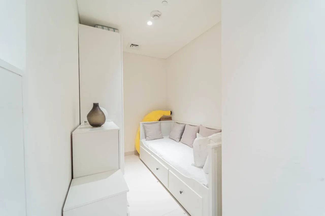 A bright bedroom with a bed, pale pillows, and a bedside cabinet.