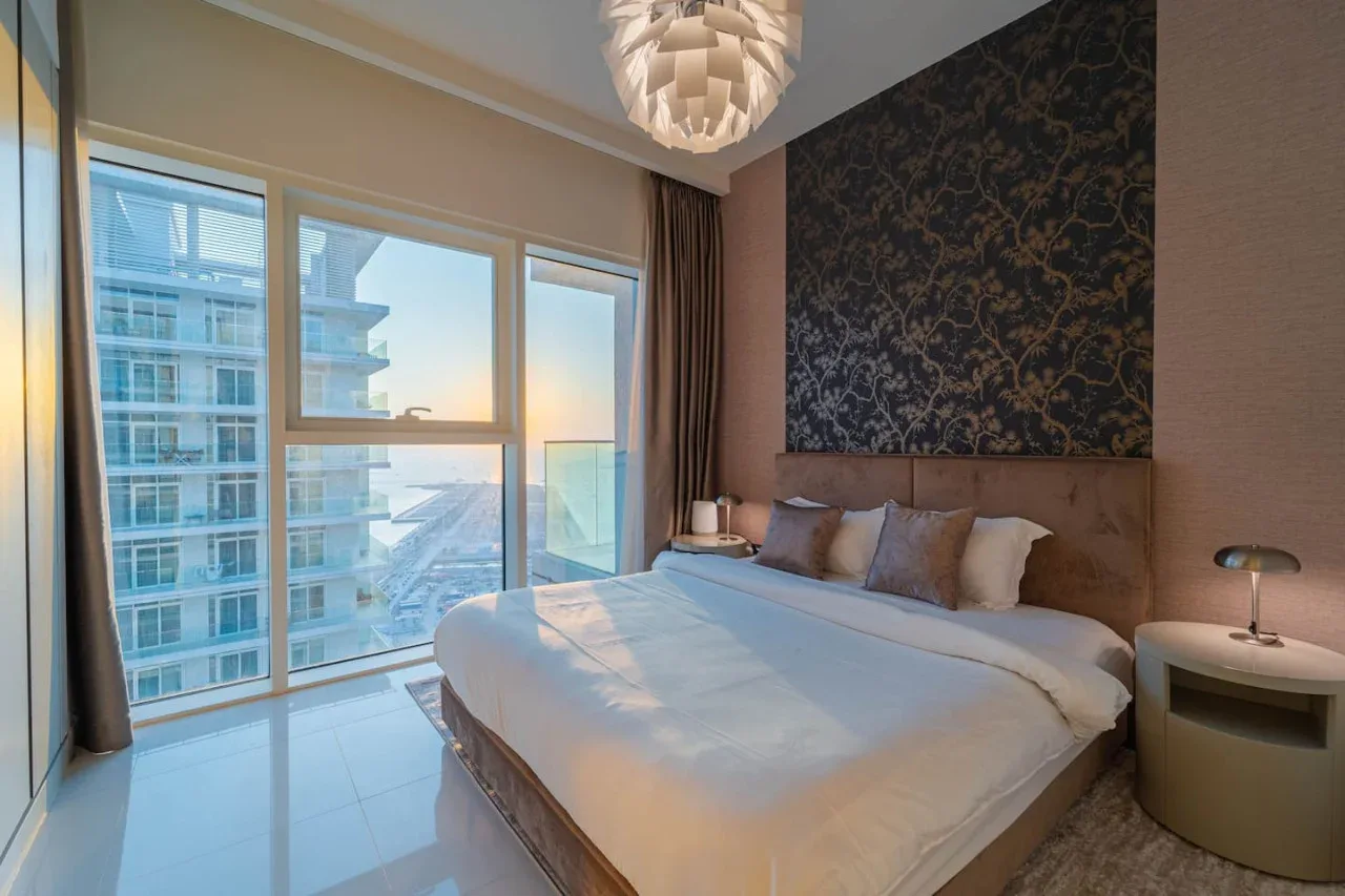 A bedroom with a bed beside large windows overlooking nearby buildings and water.