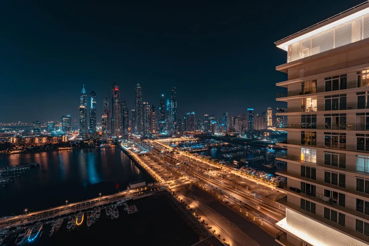 Nighttime cityscape with illuminated skyscrapers and a brightly lit highway beside the water.