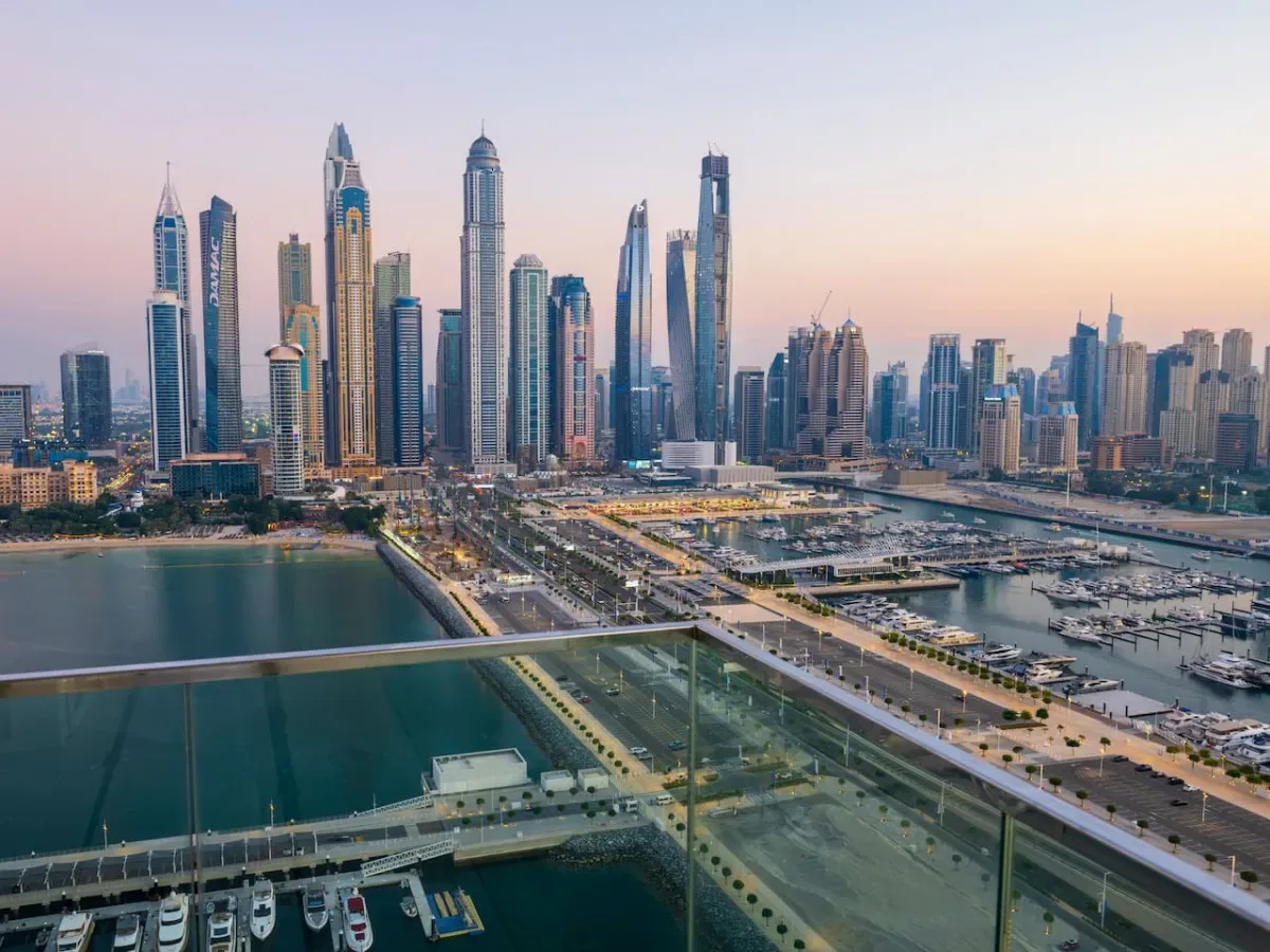 A waterfront skyline of tall buildings rises above roads and marina basins at dusk.
