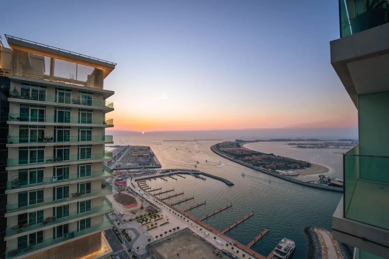 A waterfront harbor and marina at sunset, framed by modern buildings.