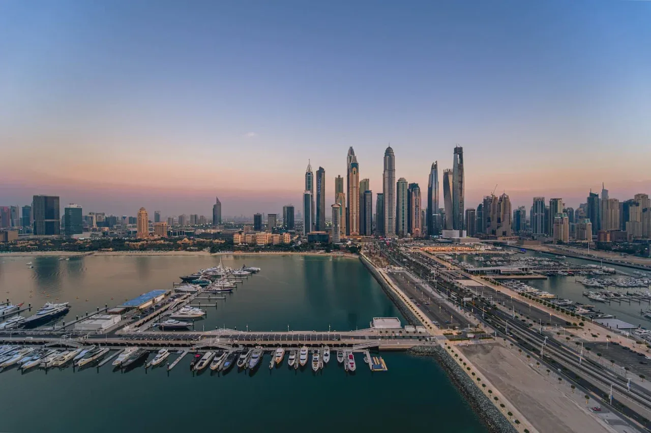 A waterfront marina and road lead toward a city skyline at dusk.