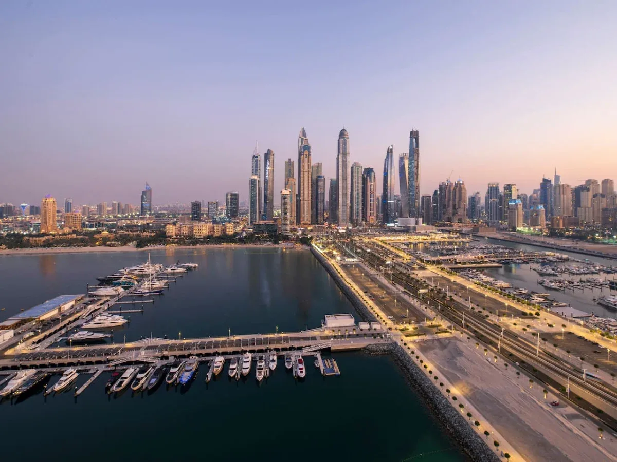 A waterfront city skyline rises beyond a marina and illuminated roads at dusk.