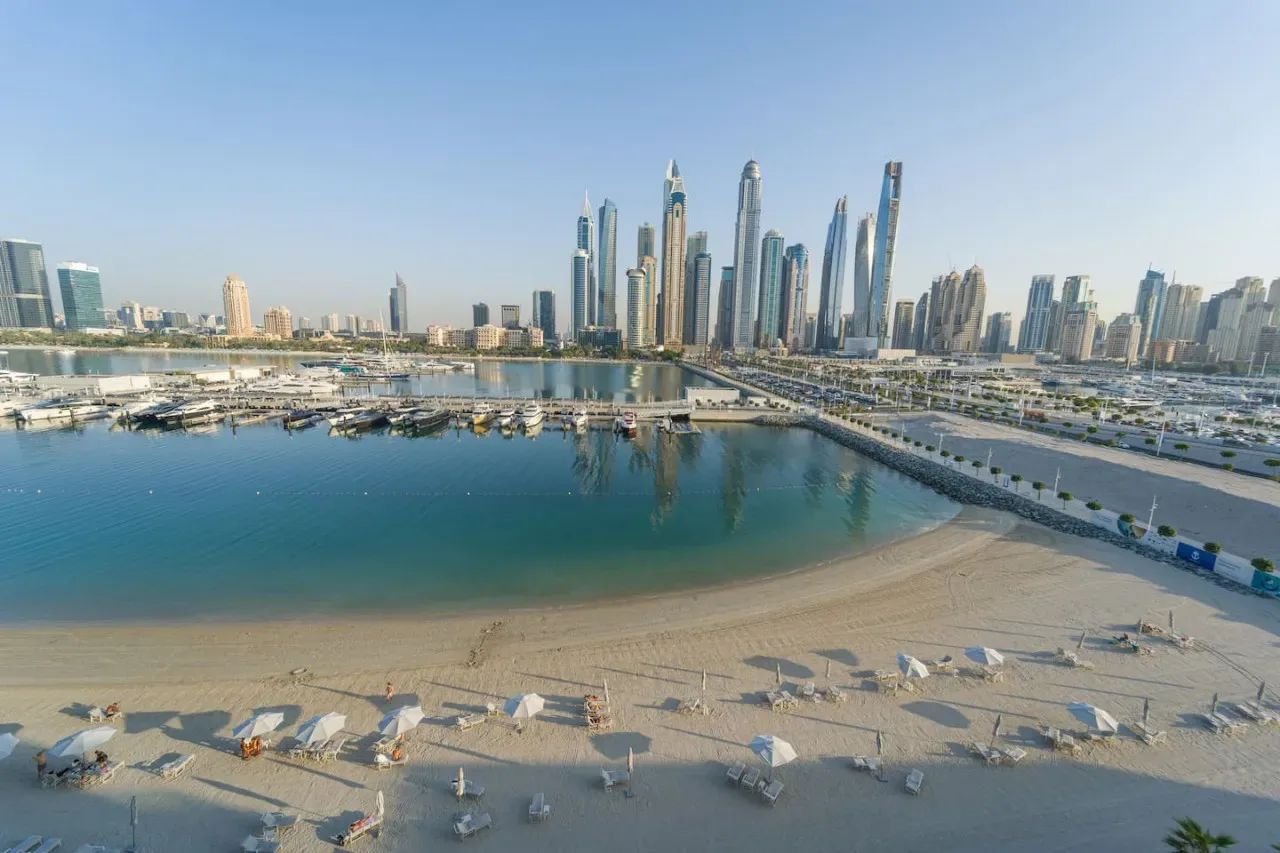 A sandy waterfront beach and marina sit below a modern city skyline.