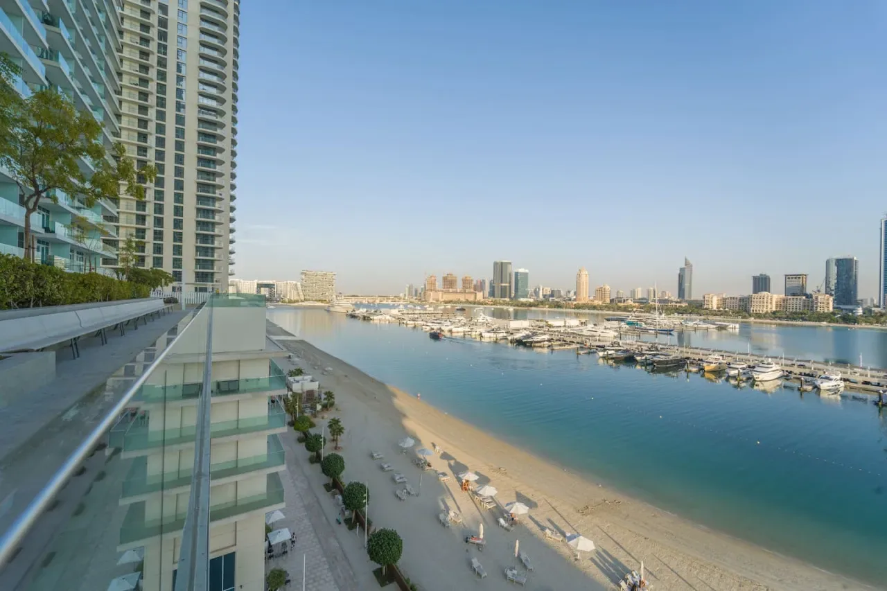 A beachfront high-rise overlooks a sandy shore, marina, and distant city skyline.