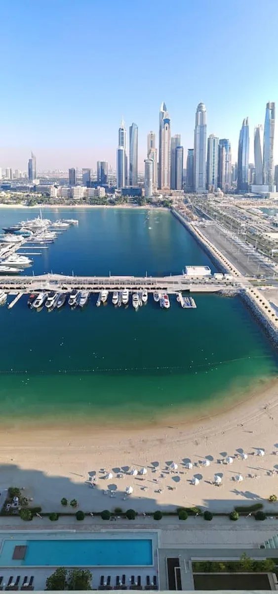 Aerial view of a marina and sandy beach with a high-rise skyline beyond.