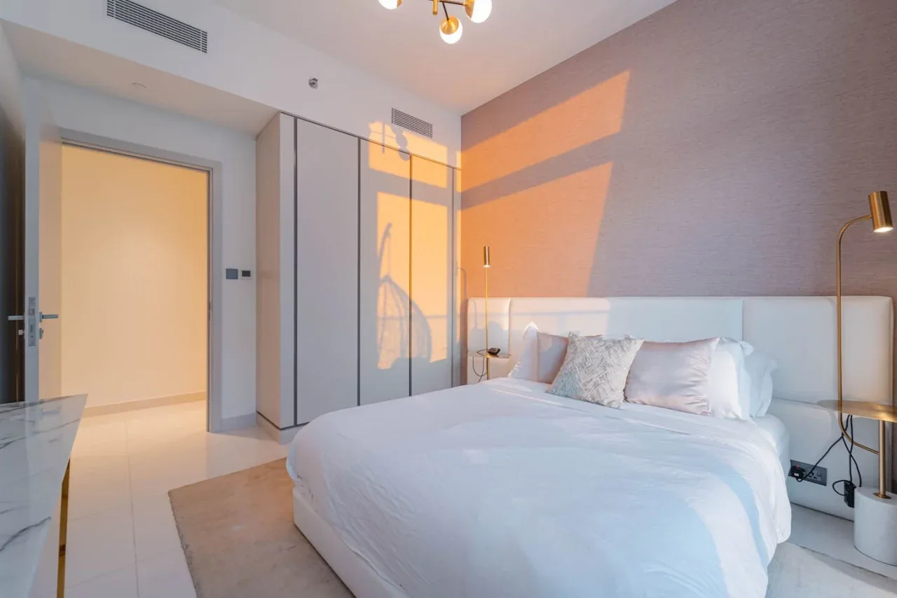 A bedroom with a large bed, pale bedding, and sunlight falling across the wall.