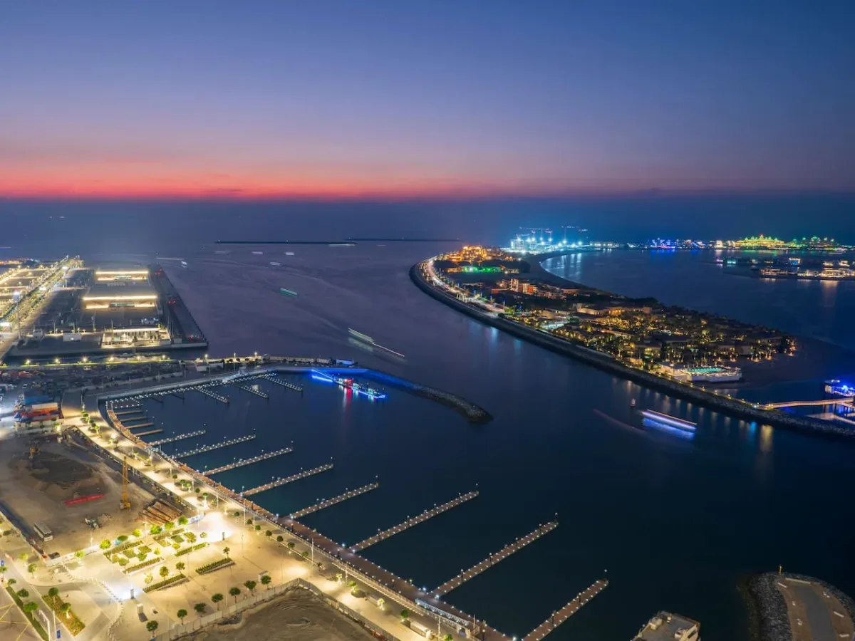 An aerial view of a waterfront harbor at dusk, with illuminated marinas along the shore.