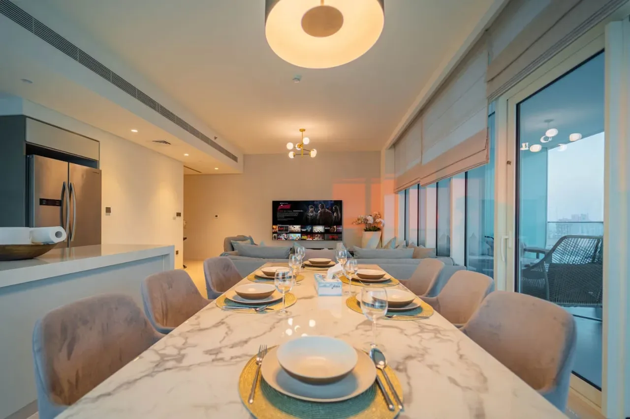 A dining area with a long table beside large windows overlooking a balcony.