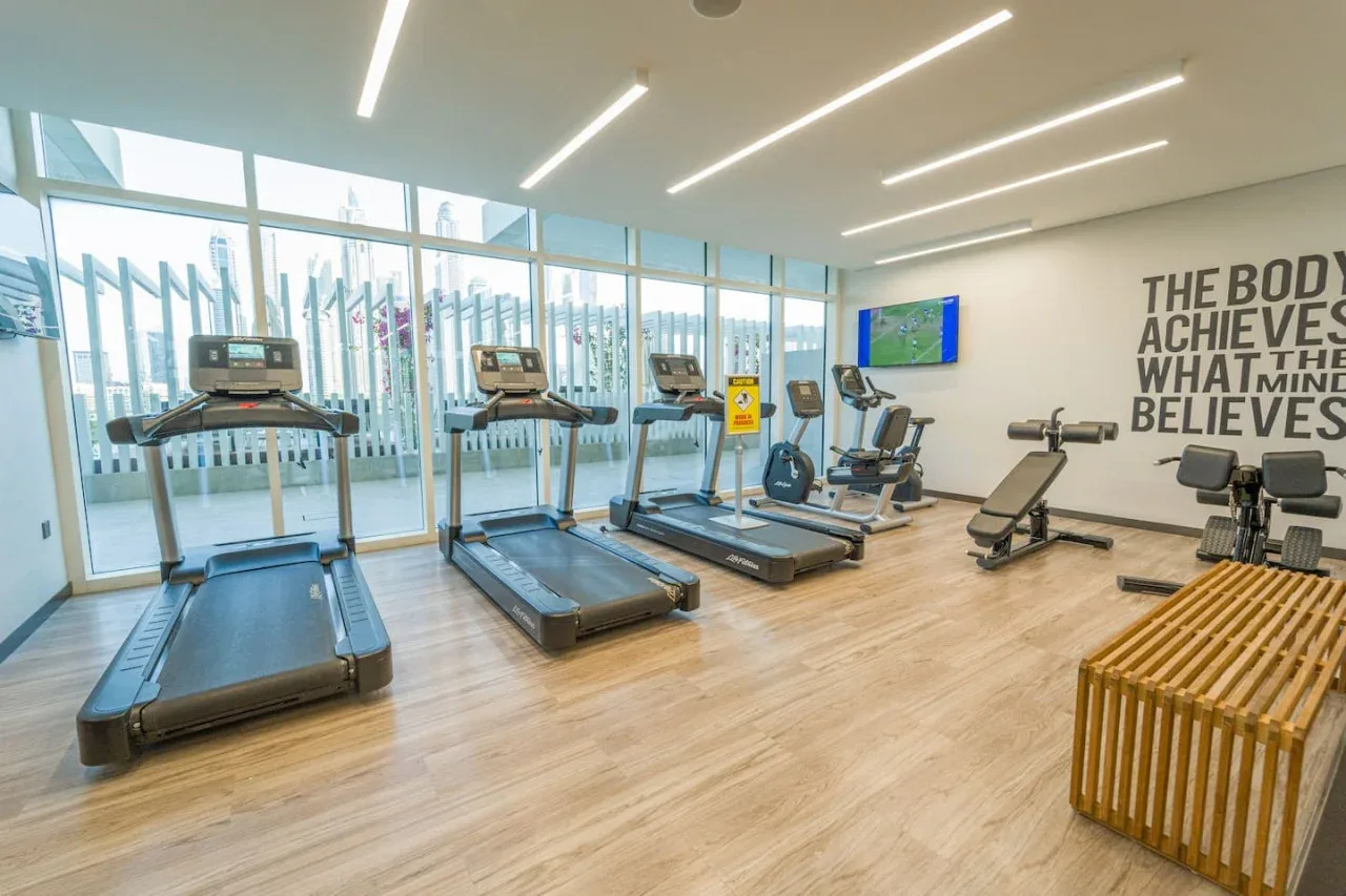 A gym with treadmills and exercise bikes beside floor-to-ceiling windows.