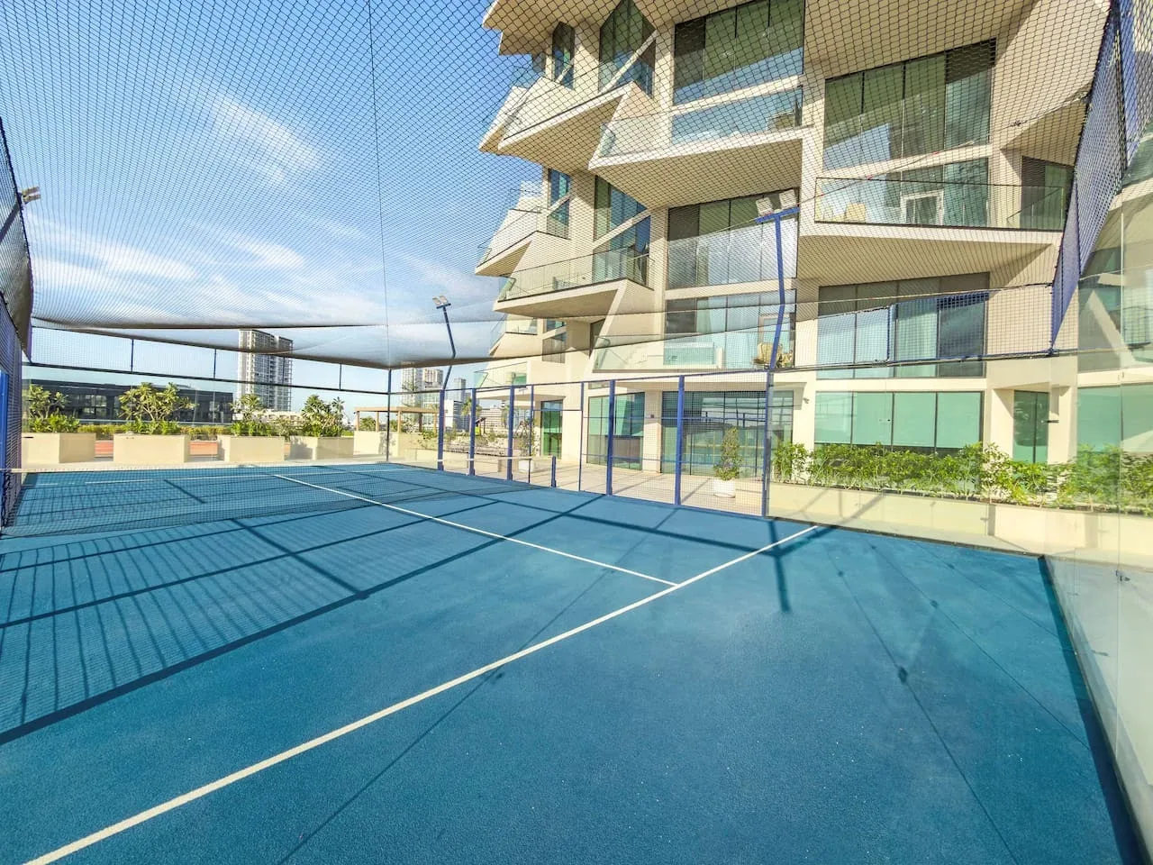 Una pista de tenis azul está rodeada por una malla junto a un edificio de varios pisos.