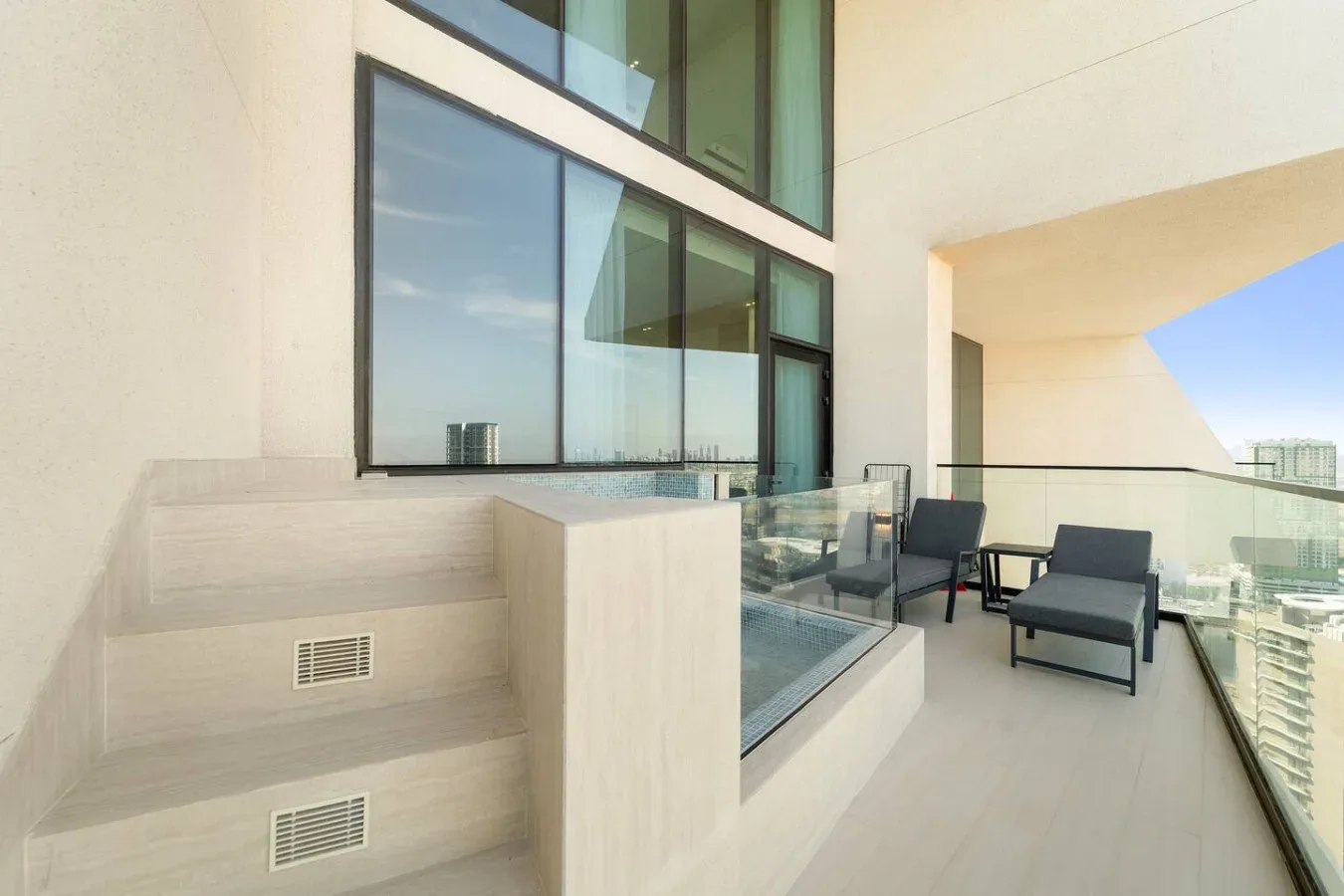 Un balcón moderno con barandillas de vidrio, asientos y grandes ventanas.