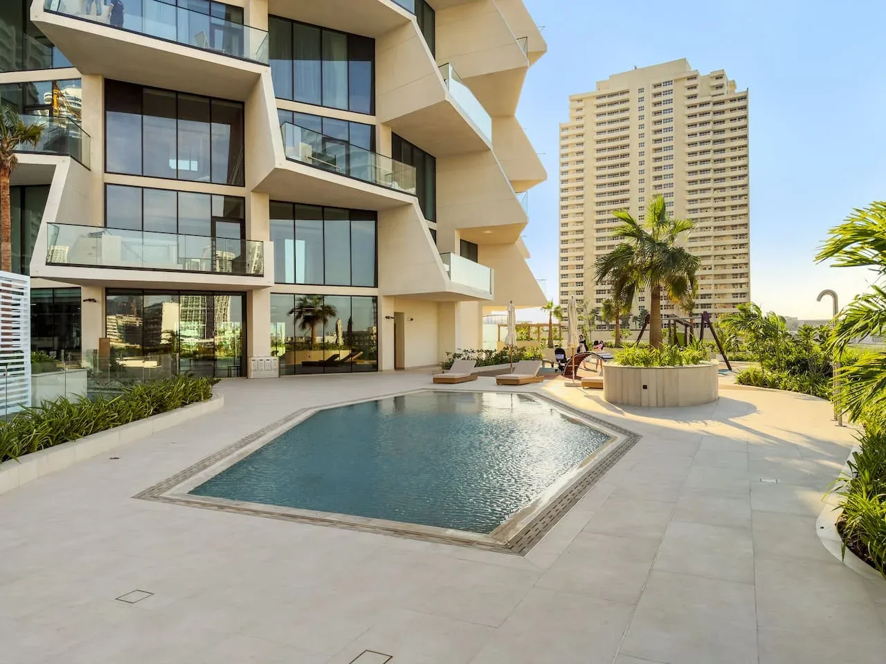 Una piscina exterior está junto a un edificio moderno con fachada de vidrio y una terraza ajardinada.