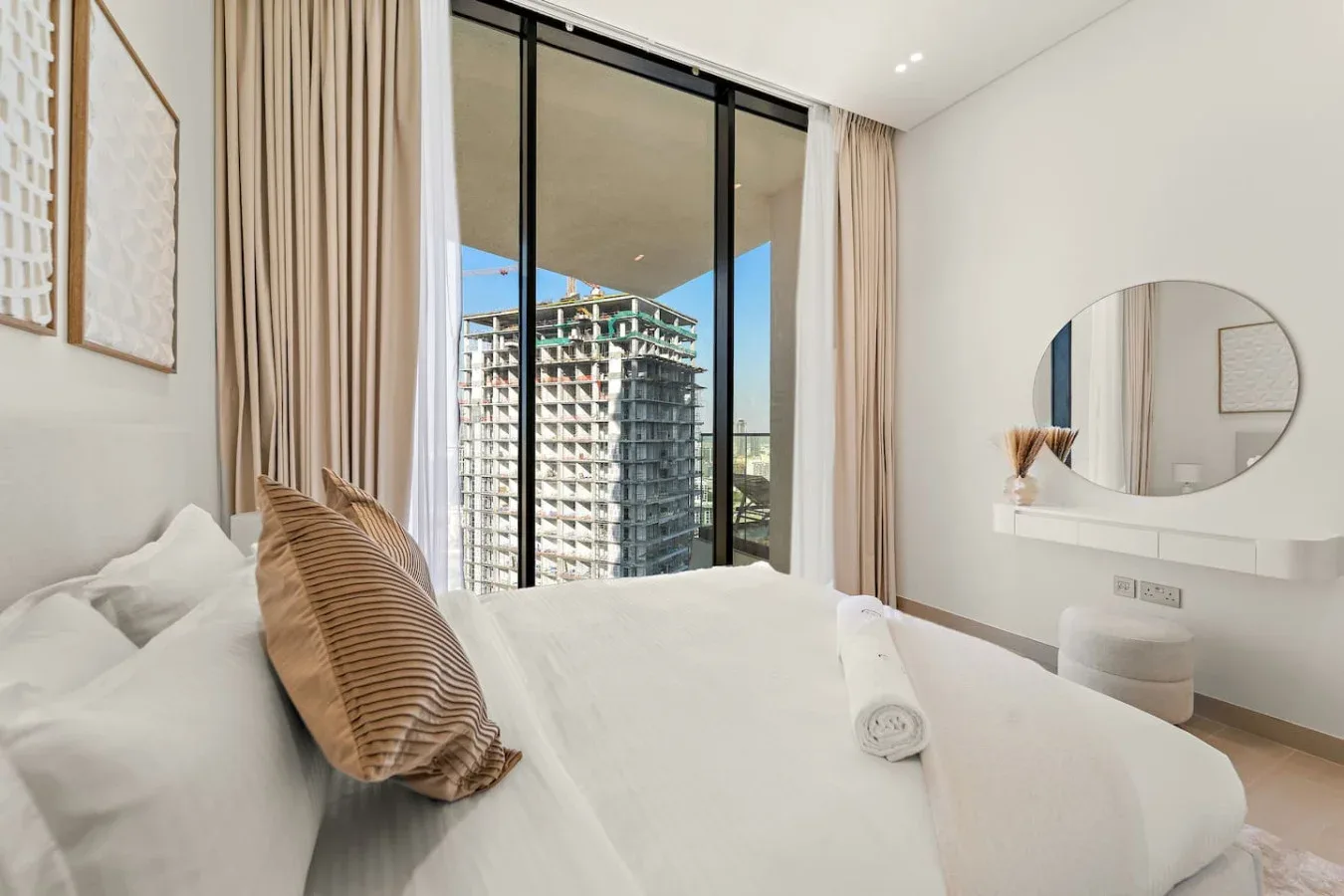Un dormitorio luminoso con una cama tendida y una gran ventana con vista a edificios cercanos.