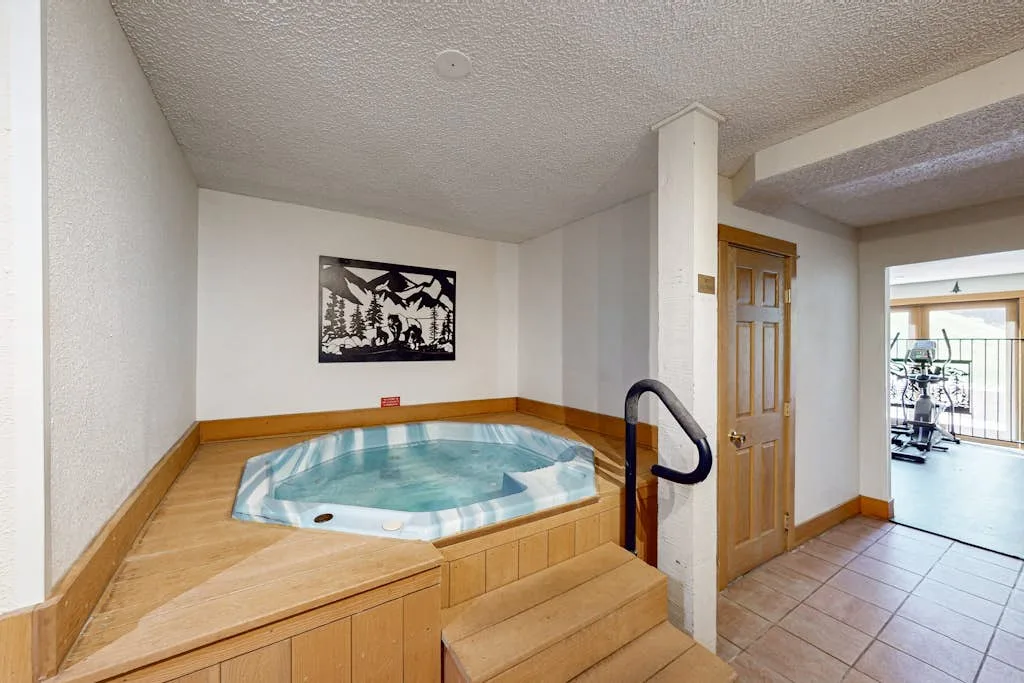 An indoor hot tub sits beside wooden steps, with a doorway opening to a bright room.